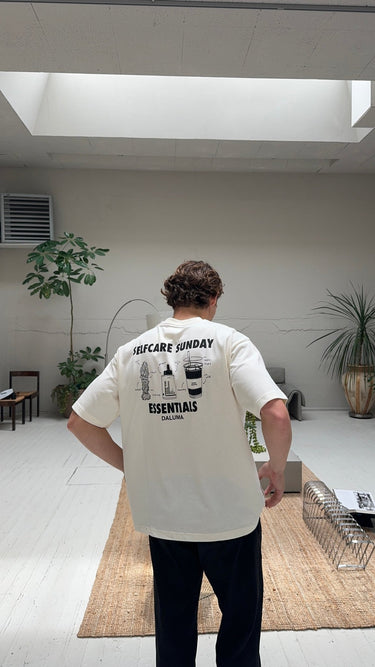 Eine Person mit lockigen braunen Haaren steht in einem Innenraum auf einem hellen Holzboden und blickt in dem übergroßen JOSEPH ATELIER X DALUMA SELFCARE SUNDAY TEE von DALUMA in die Ferne. Der Raum ist mit modernen Möbeln, Zimmerpflanzen und neutralem Dekor ausgestattet.