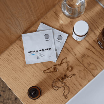 Zwei Päckchen DALUMA DEEP HYDRATION FEUCHTIGKEITSMASKE, ein kleiner Krug, eine goldene Kette und ein Glas ruhen auf Holz neben einem weißen Waschbecken - ideal für den Genuss von Tiefenhydration mit natürlichen Zutaten.