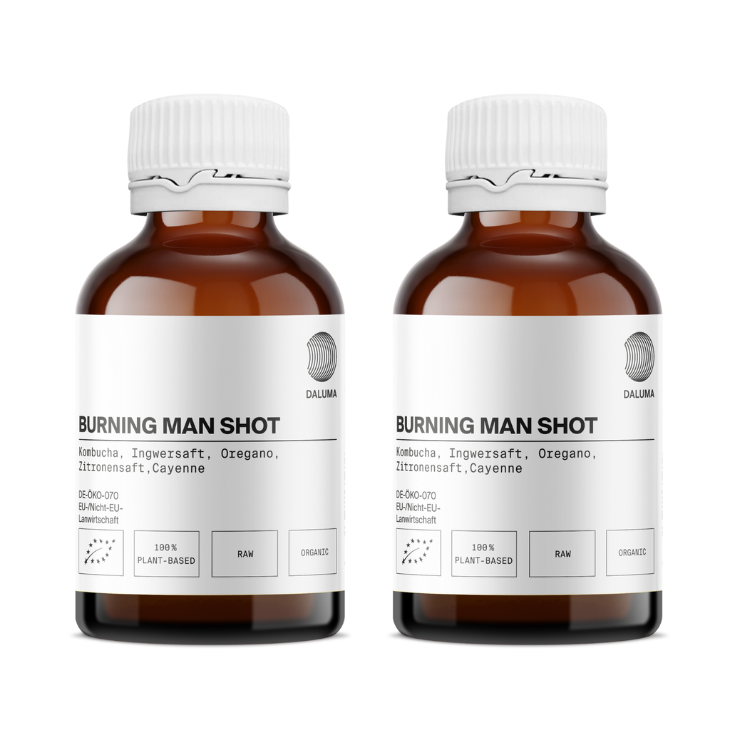 Zwei braune Glasflaschen DALUMA BURNING MAN INGWERSHOT mit weißem Deckel, mit Kombucha, Ingwer, Oregano, Zitronensaft und Cayenne. Nebeneinander abgebildet - perfekt zur Stärkung des Immunsystems. Erhältlich in einer 24er-Packung.