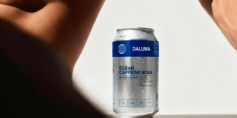 Eine kalte, mit Wassertropfen bedeckte Dose Daluma Clean Caffeine BCAA Drink steht aufrecht auf einer weißen Fläche zwischen zwei unscharfen, gebräunten Armen oder Beinen. Der Hintergrund ist hell beleuchtet.
