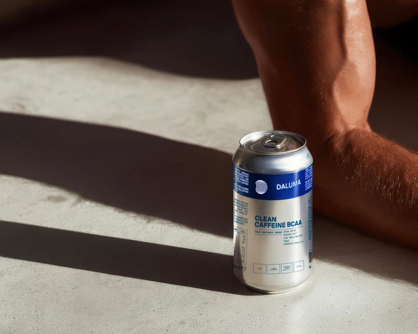 Eine Dose mit der Aufschrift DALUMA ENERGY & STRENGTH DUO ruht auf einer sonnenbeschienenen Fläche in der Nähe des nackten Arms und Beins einer Person und wirft deutliche Schatten auf den Boden.