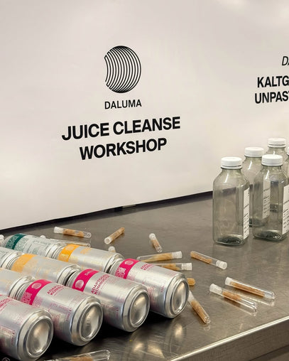 Mehrere Dosen, Glasflaschen und kleine Reagenzgläser sind auf einem Metalltisch vor einem Schild mit der Aufschrift "DALUMA JUICE CLEANSE WORKSHOP" angeordnet. Die Szene deutet auf eine Werkstatt für Säfte oder Gesundheitsgetränke hin.
