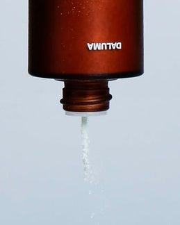 Eine braune Flasche mit der Aufschrift DALUMA, die auf dem Kopf steht, spendet ein feines weißes Pulver von GREEN COFFEE CLEANSER ENZYMPEELING für die exfolierende Gesichtspflege aus ihrer offenen Düse vor einem schlichten Hintergrund.