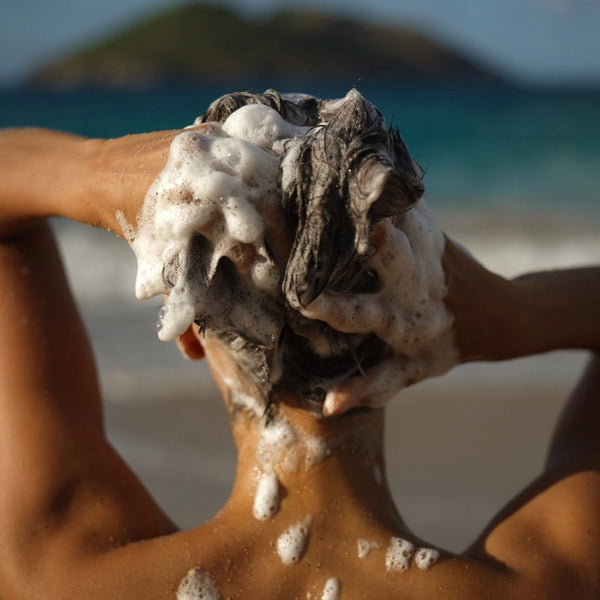 Eine Person steht am Strand, von hinten gesehen, und wäscht sich die Haare mit Shampoo, wobei dicker, weißer Schaum entsteht, der ihre Haare und Hände bedeckt. Im Hintergrund sind das Meer und eine entfernte Insel zu sehen.