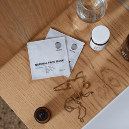 Zwei weiße Päckchen mit der Aufschrift HYDRATING ESSENTIALS by DALUMA stehen auf einer Holzfläche neben einer Halskette, einem kleinen Glas und einer braunen Flasche in einem Raum, der wie ein Badezimmer oder ein Waschbecken aussieht.