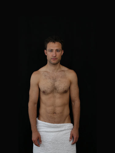 Ein hemdsärmeliger Mann steht mit dem Gesicht nach vorne, in ein weißes Handtuch um die Taille gewickelt, vor einem soliden schwarzen Hintergrund.