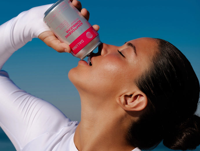 Eine Frau mit weißem Oberteil trinkt vor einem strahlend blauen Himmel aus einer silber-rosa Dose, wobei sie den Kopf zurücklehnt und die Dose über den Mund hält.