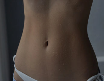 Nahaufnahme des nackten Mittelteils einer Person, die ihren Bauch und einen Teil der weißen Unterwäsche zeigt. Auf der Haut sind Wassertropfen zu sehen, was darauf hindeutet, dass sie gerade trainiert oder geduscht hat.