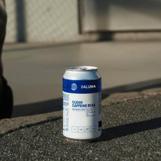 Eine DALUMA Clean Caffeine BCAA-Getränkedose steht aufrecht auf einer rauen Außenfläche, wobei das Sonnenlicht Schatten wirft und der Hintergrund mit einem Zaun und einem Bürgersteig unscharf ist.