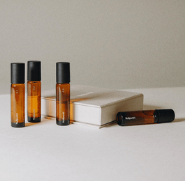 Die Nullpunkt Complete Collection besteht aus vier Braunglas-Rollerflaschen mit schwarzen Verschlüssen, die neben einem geschlossenen beigen Buch auf einem minimalistischen, neutralen Hintergrund angeordnet sind.