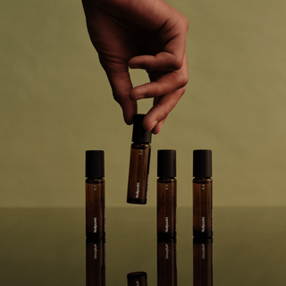 Eine Hand hält eine bernsteinfarbene Rollflasche der NULLPUNKT COMPLETE COLLECTION von Nullpunkt über einer reflektierenden Oberfläche. Drei passende Flaschen und minimalistische Etiketten stehen vor einem Hintergrund in gedämpftem Grün für einen eleganten, natürlichen Look.