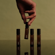 Eine Hand hält eine von vier NULLPUNKT COMPLETE COLLECTION Braunglas-Rollerflaschen von Nullpunkt mit schwarzer Kappe, die auf einer spiegelnden Fläche vor olivgrünem Hintergrund angeordnet sind.