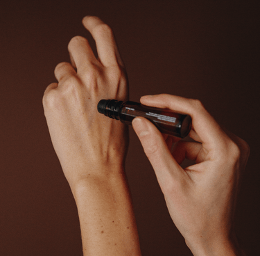 Jemand trägt das nährende Serum von Nullpunkt aus der NULLPUNKT COMPLETE COLLECTION mit einem dunkelbraunen Rollerbehälter auf, der vor einem warmen braunen Hintergrund steht.