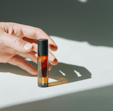 Eine Hand greift nach der Braunglas-Apothekerflasche Nullpunkt Grade 0.1 Relieve mit schwarzem Verschluss und wirft einen Schatten auf eine weiße Fläche in hellem, natürlichem Licht.