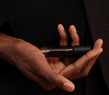 Eine Person in einem dunklen Anzug hält einen braunen Glasflakon des Parfums Nullpunkt GRADE 0.3 REFOCUS in der Hand, wobei ihre Finger den Duft von Nullpunkt vor einem schattigen Hintergrund sanft umschmeicheln.