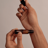 Eine Person hält einen kleinen Roll-on-Flakon aus Braunglas mit der Aufschrift NULLPUNKT GRADE 0.3 REFOCUS von Nullpunkt in der Hand und schraubt den schwarzen Verschluss mit einer Hand ab, während sie den Flakon vor einem minimalistischen beigen Hintergrund hält.