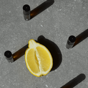 Eine halbierte Zitrone auf einer grauen Fläche ist von vier braunen Nullpunkt NULLPUNKT GRADE 0.4 REFUEL-Flaschen mit schwarzen Verschlüssen umgeben, die jeweils lange Schatten werfen.