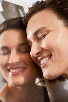 Eine Person mit kurzen braunen Haaren lächelt, ihre Wange gegen einen Spiegel gerichtet, und hebt die glatte Haut und die Vorteile von DALUMA PREBIOTIC PURIFYING GESICHTSSERUM für eine gesunde Hautbarriere hervor.