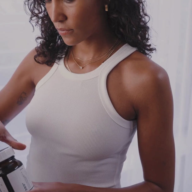 Eine Frau mit lockigem Haar und Goldschmuck öffnet eine Flasche Daluma 360 DAILY ESSENTIALS SUPPLEMENT DOUBLE. Sie trägt ein weißes geripptes ärmelloses Oberteil vor einem sanft beleuchteten weißen Hintergrund.