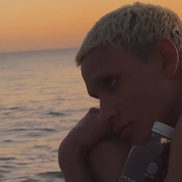 Eine Person mit kurzem, hellem Haar sitzt bei Sonnenuntergang am ruhigen Meer und hält eine klare Flasche Daluma BETTER GUT SAFTKUR 3 TAGE in der Hand, während der Himmel im Hintergrund orange leuchtet.