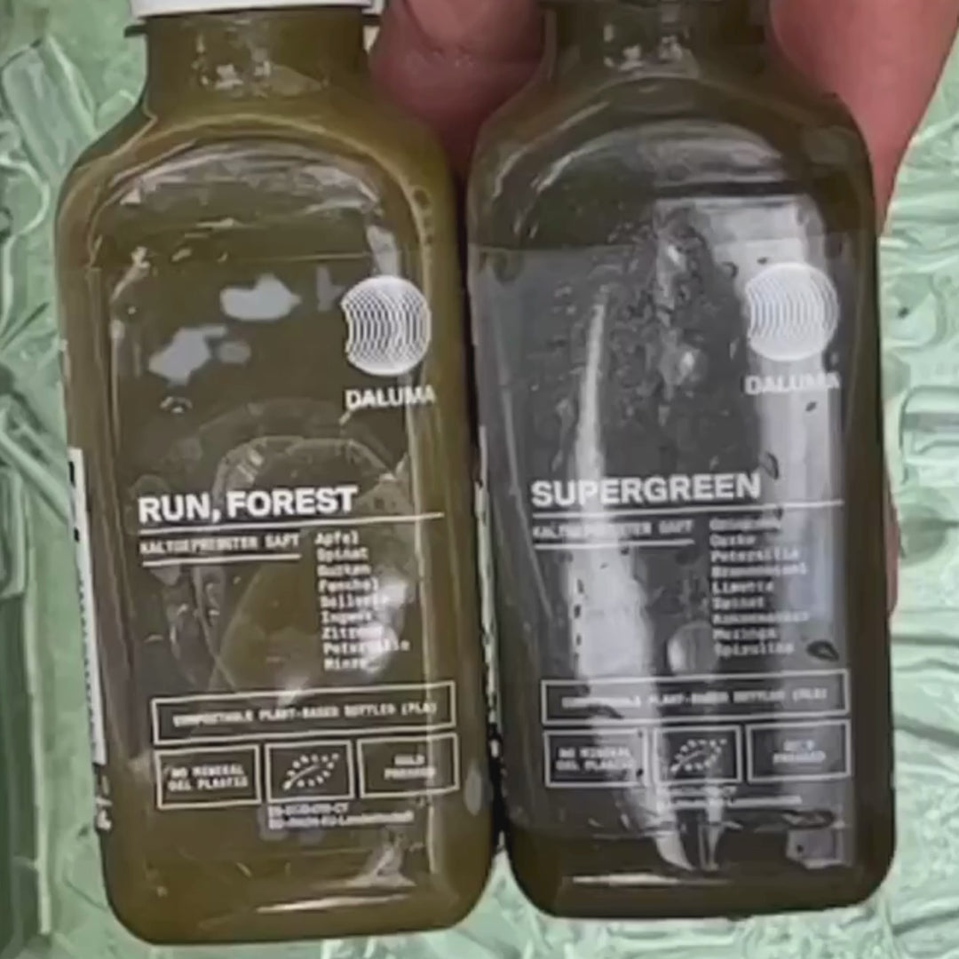 Zwei grüne Saftflaschen der Marke DALUMA mit den Aufschriften RUN, FOREST und SUPERGREEN werden nebeneinander gehalten. Auf beiden Flaschen sind die Zutaten aufgelistet und die Symbole der Bio-Zertifizierung abgebildet.