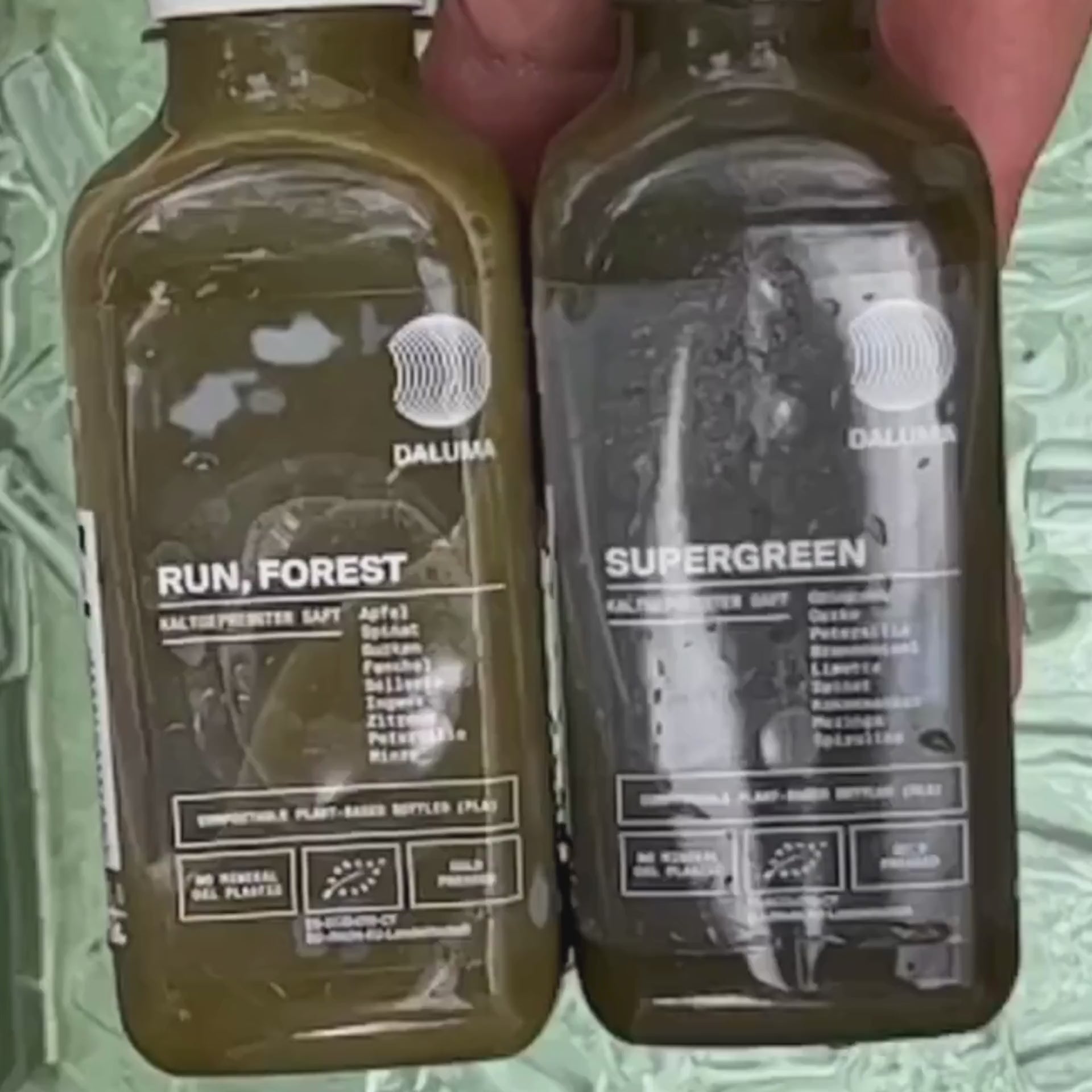 Zwei grüne Saftflaschen der Marke DALUMA mit den Aufschriften RUN, FOREST und SUPERGREEN werden nebeneinander gehalten. Auf beiden Flaschen sind die Zutaten aufgelistet und die Symbole der Bio-Zertifizierung abgebildet.