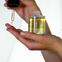 Eine Person hält eine Glasflasche mit DALUMA RADIANT GLAZE GESICHTSÖL in der einen Hand und gibt mit einer Pipette Tropfen auf ihre andere Handfläche. Der Hintergrund ist hell und schlicht.