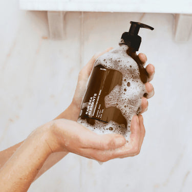 Eine Person hält einen dunkelbraunen Daluma-shop BODY CARE DUO-Spender mit schwarzer Pumpe in ihren seifigen Händen, während sie in einer Dusche vor einer hellen Kachelwand steht.