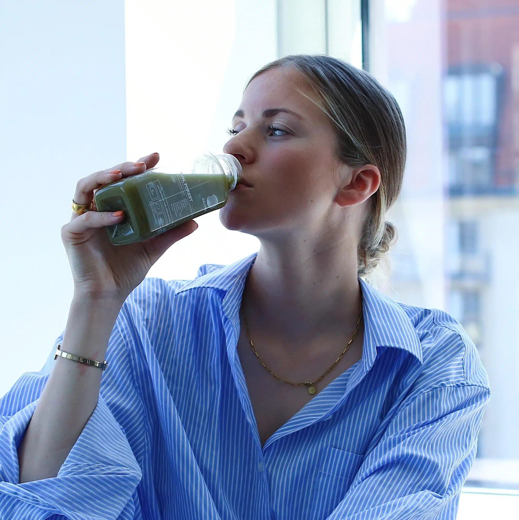 Eine Frau in einem blau gestreiften Hemd genießt DALUMA GREEN JUICE PLUS SAFTKUR 5 TAGE aus einer Glasflasche am Fenster und lässt sich die frischen Bio-Gemüsesäfte schmecken.
