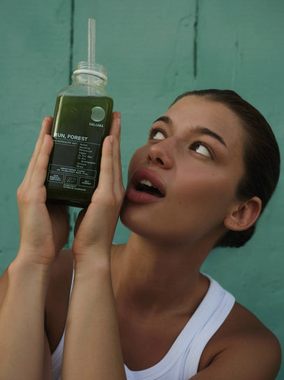 Eine Frau in einem weißen Tank-Top hält eine grüne Saftflasche mit einem Strohhalm und schaut sie überrascht oder bewundernd an, während sie vor einer grünen Wand steht.