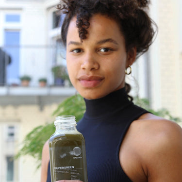 Eine Person mit lockigem Haar in einem ärmellosen schwarzen Rollkragenpullover hält eine durchsichtige Flasche DALUMA SUPERGREEN GREEN JUICE 6 PACK, während sie draußen vor Gebäuden im Hintergrund steht.