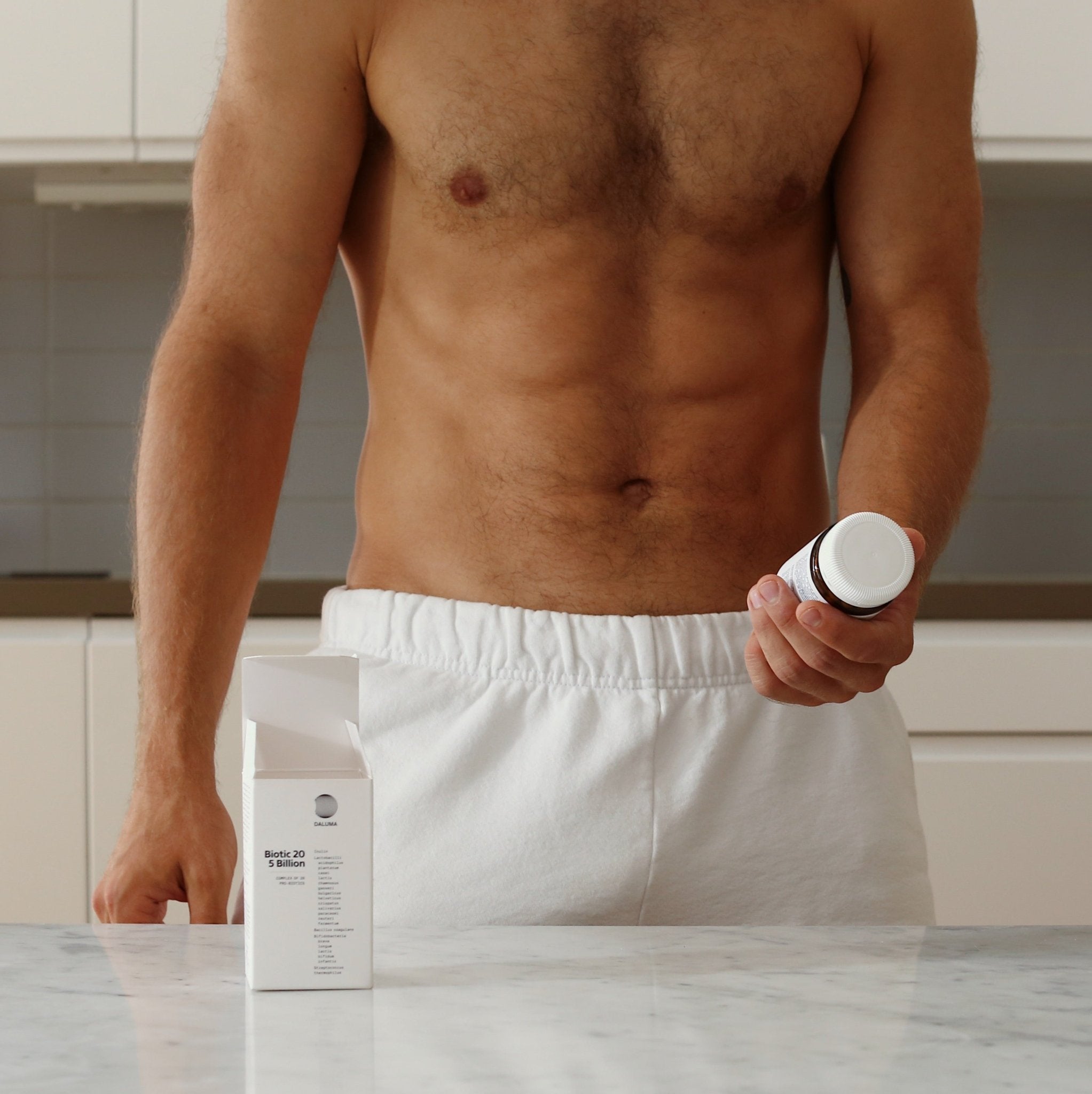 Eine hemdlose Person in weißen Shorts steht in einer Küche und hält eine SYM-BIOTIC 23/100 BILLION DARMBAKTERIEN DOUBLE Ergänzungsflasche von DALUMA in der Hand. Auf dem Tresen steht eine offene Schachtel mit magensaftresistenten Kapseln. Ihr Gesicht ist nicht zu sehen.