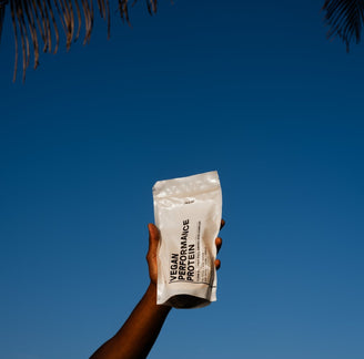 Eine Hand hält einen weißen DALUMA-Beutel mit der Aufschrift VEGANES PERFORMANCE PROTEIN NEUTRAL vor einem klaren blauen Himmel mit Palmenblättern am oberen Bildrand hoch.
