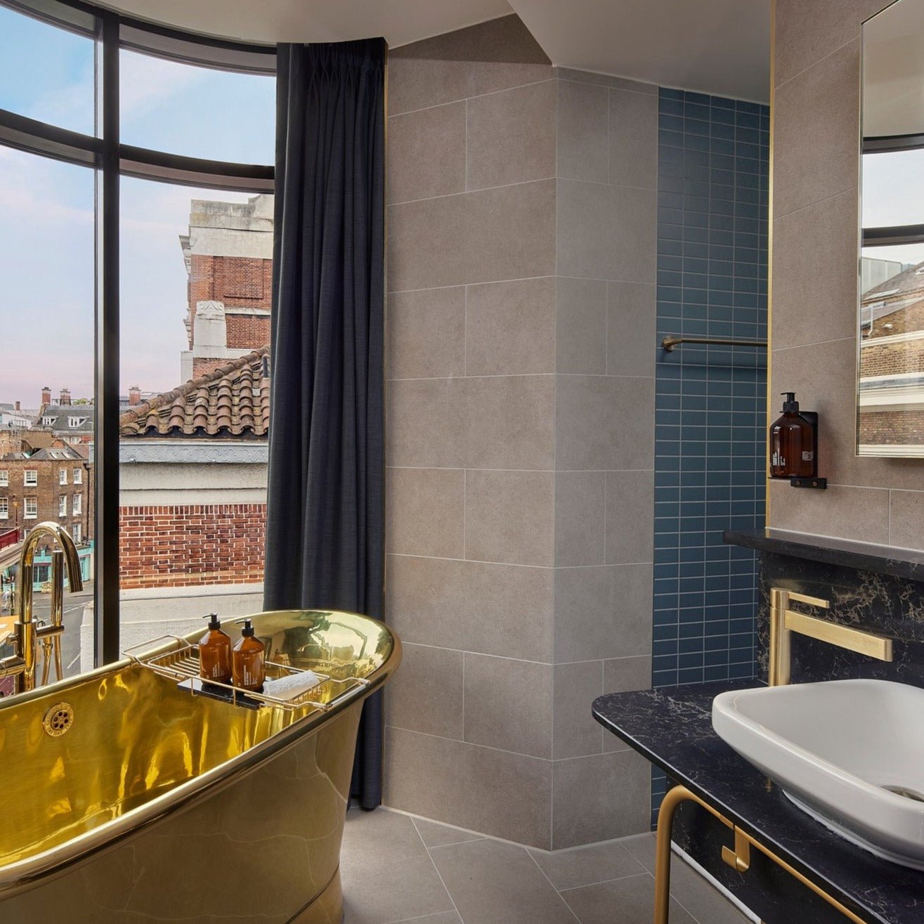 Ein modernes Badezimmer mit der BODYCARE WANDHALTERUNG von DALUMA, einer freistehenden goldenen Wanne an einem geschwungenen Fenster, einer schwarzen Arbeitsplatte mit weißem Waschbecken und hellgrauen Fliesen. Durch das Fenster sind blaue Fliesen und freiliegende Ziegel zu sehen.